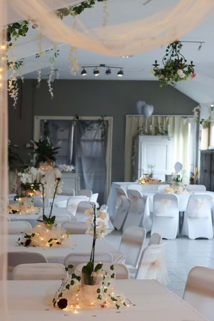 An elegantly decorated event space featuring floral arrangements with white orchids and dark red roses, placed on tables wrapped in white tablecloths. Each table is adorned with warm string lights int