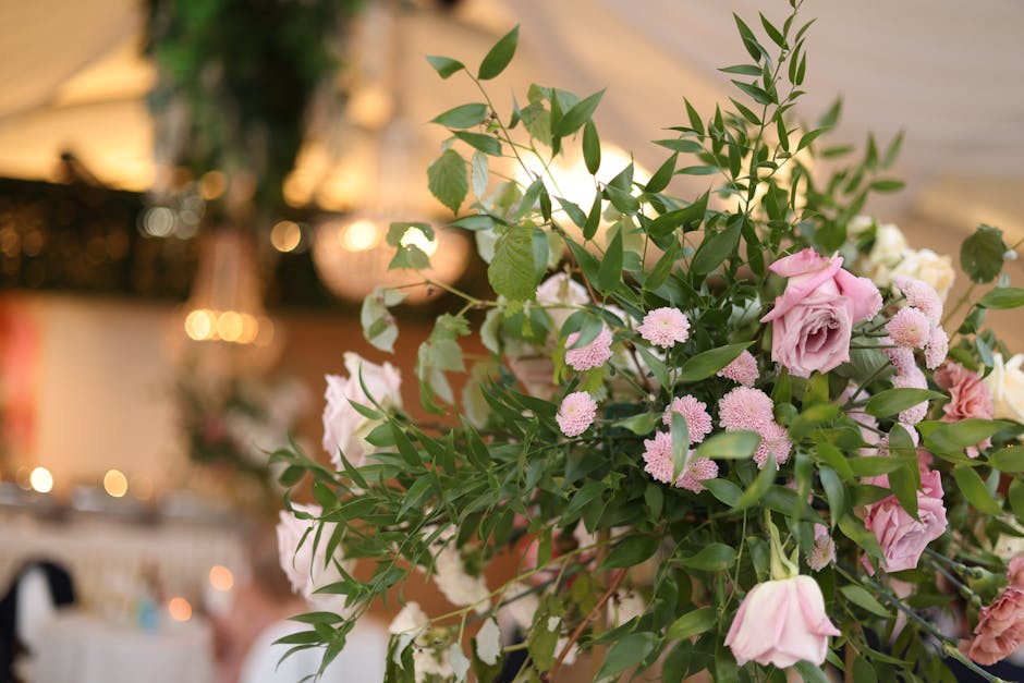 A lush floral arrangement featuring light pink roses and small, round pink chrysanthemums, accented with abundant green foliage including long, slender leaves and rounded herbaceous stems. The bouquet
