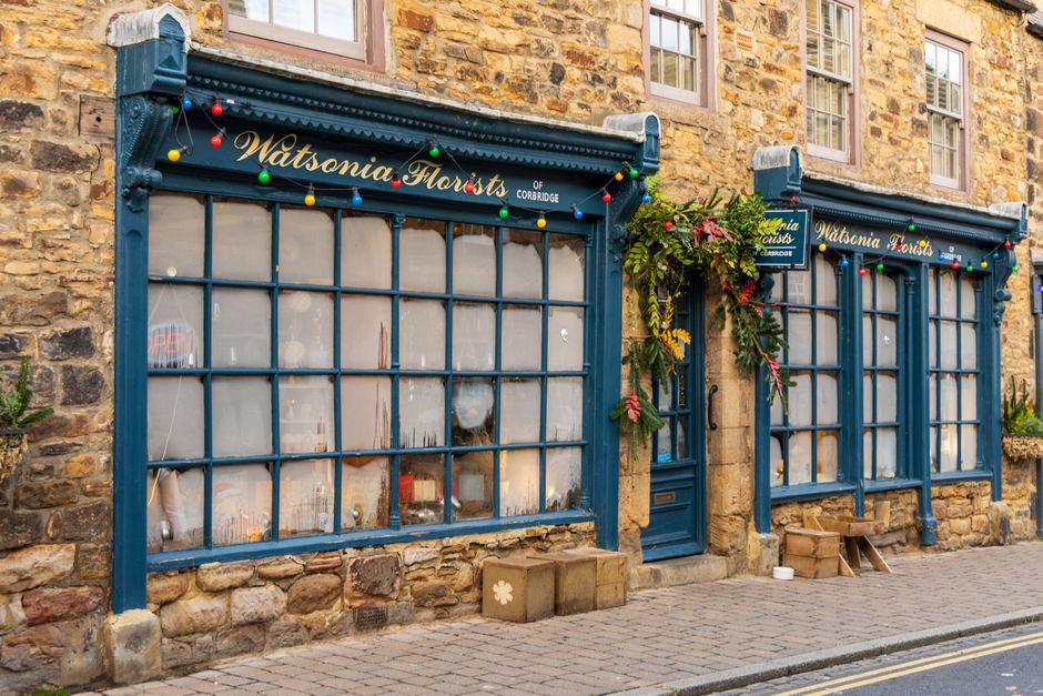 A floral arrangement with various fresh flowers, including pink, yellow, and green blooms, is positioned around the blue entrance door of Watsonia Florists of Corbridge. The arrangement features lush 
