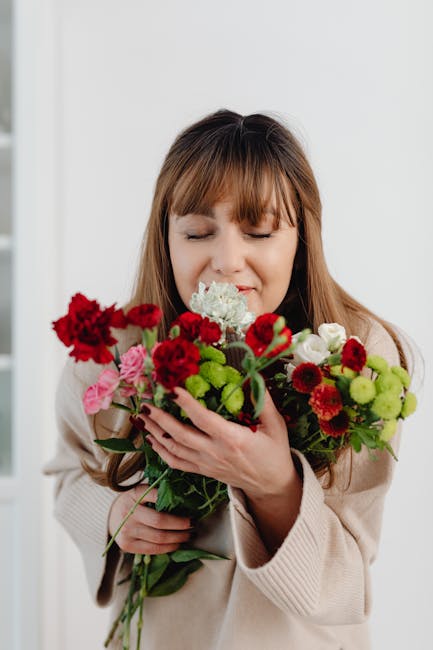 A woman with light brown hair, smiling softly with her eyes closed, gently holding a vibrant bouquet of fresh flowers close to her face. The bouquet features a variety of blooms including deep red car