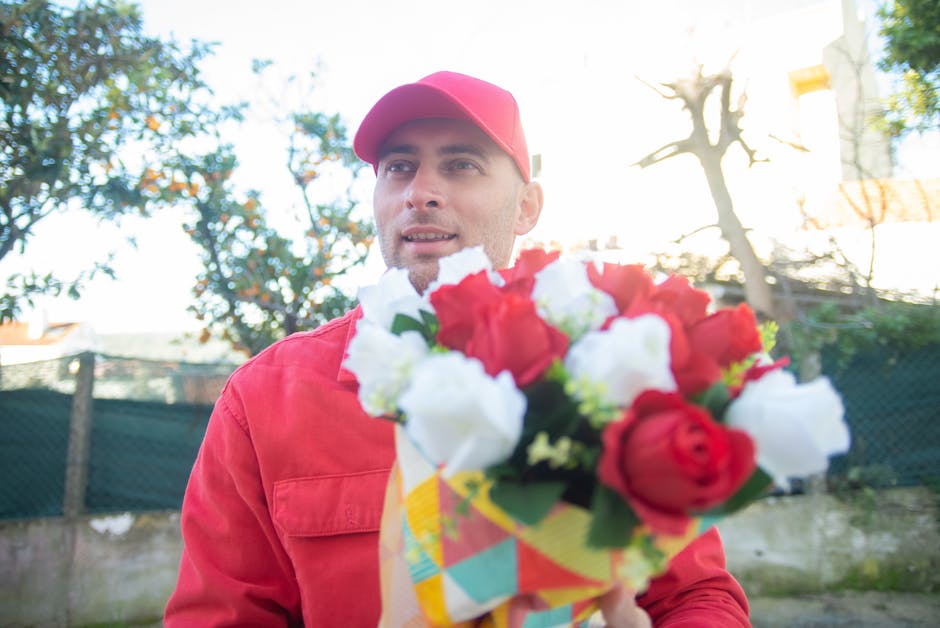 A man wearing a red cap and red jacket is holding a vibrant bouquet of fresh flowers, including red roses and white blooms, wrapped in colorful paper. He is outdoors, with sunlight filtering through t