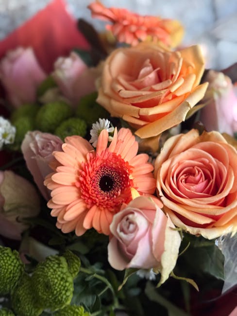 A close-up view of a vibrant floral bouquet arranged with peach and blush pink roses, complemented by soft pink spray roses and coral-orange gerbera daisies. The bouquet is wrapped in pink tissue pape