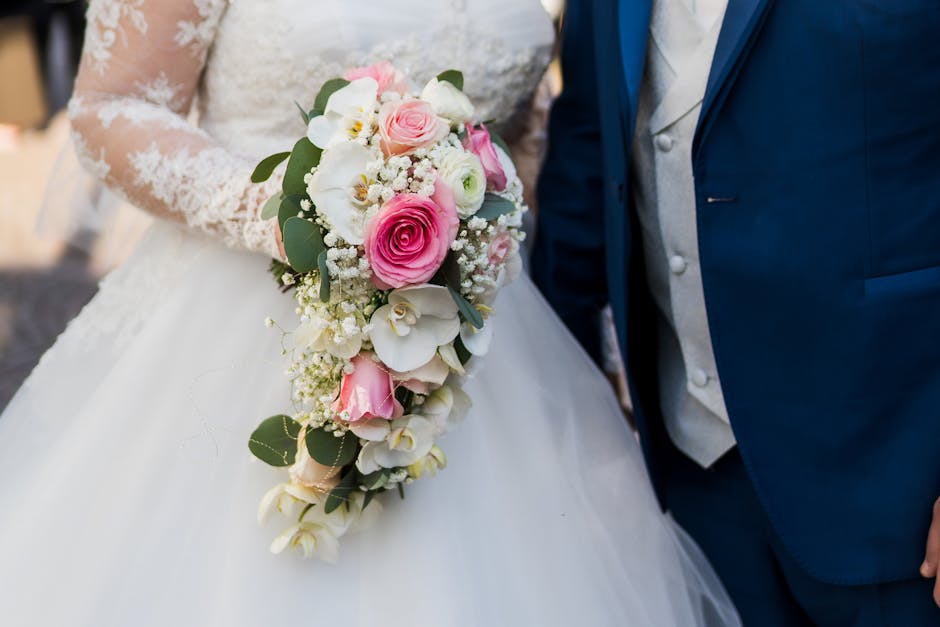 A close-up of a bride holding a cascading floral bouquet featuring pink roses, white orchids, and baby's breath, accented with green eucalyptus leaves, resting against her white lace wedding gown. To 